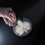 A hand holds a round black ESCLUSSO pendant featuring a detailed white orchid flower embedded in scented Wax, set against a dark black background.