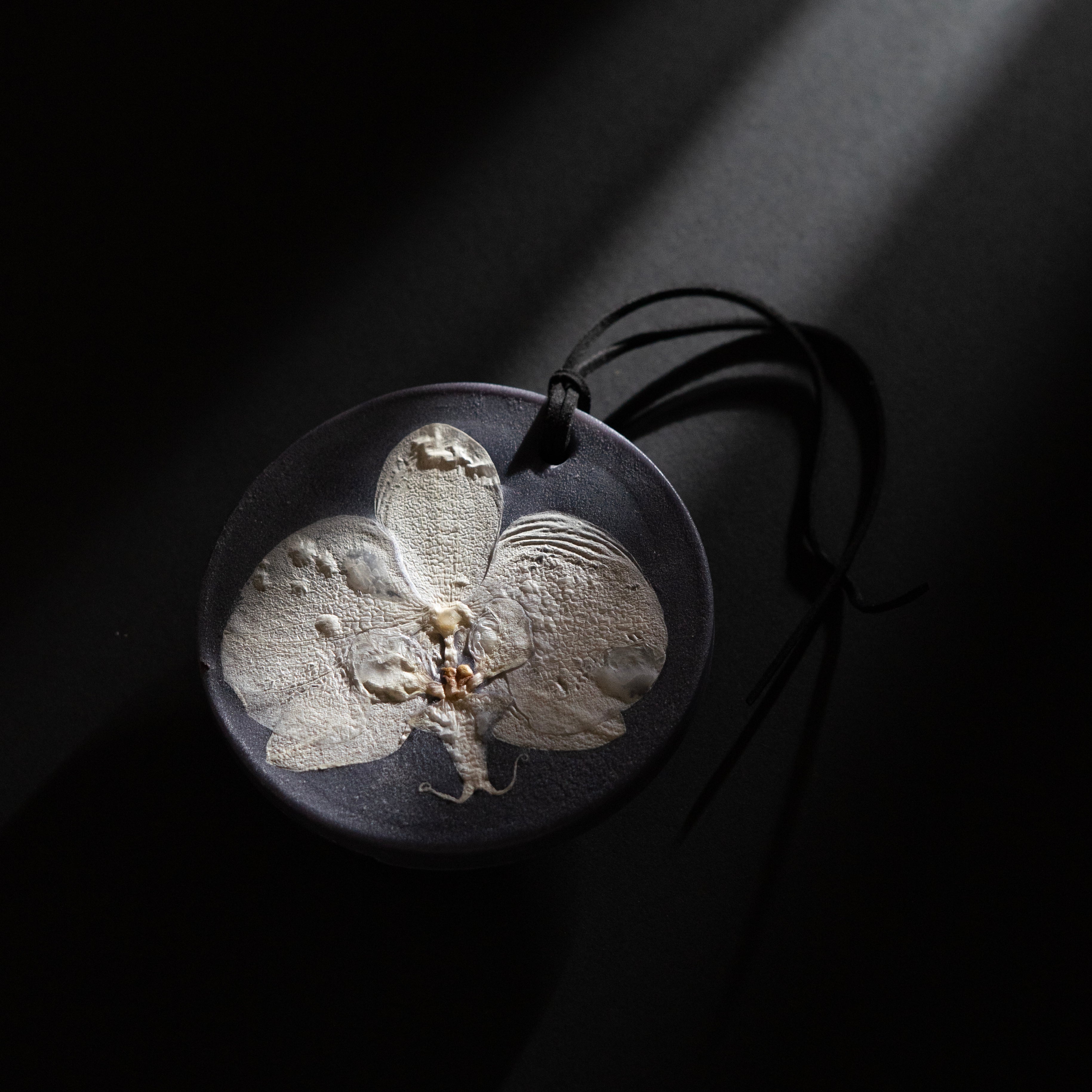 ESCLUSSO ORCHID NOCTURNE scented botanical wax sachet featuring a white orchid preserved in dark charcoal wax with black hanging cord.